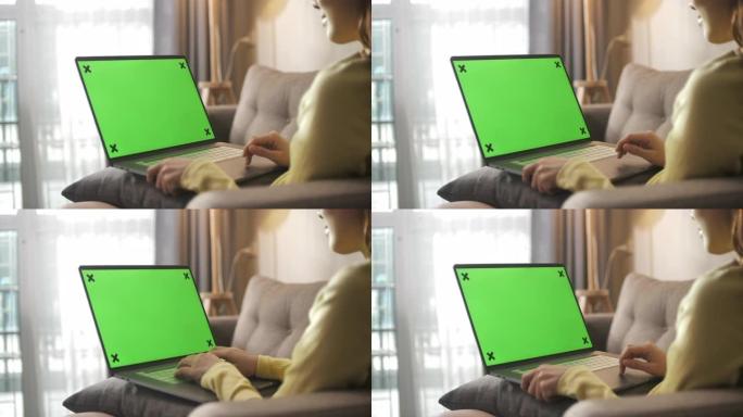 亚洲妇女坐在沙发上，在家中使用带有绿屏的笔记本电脑