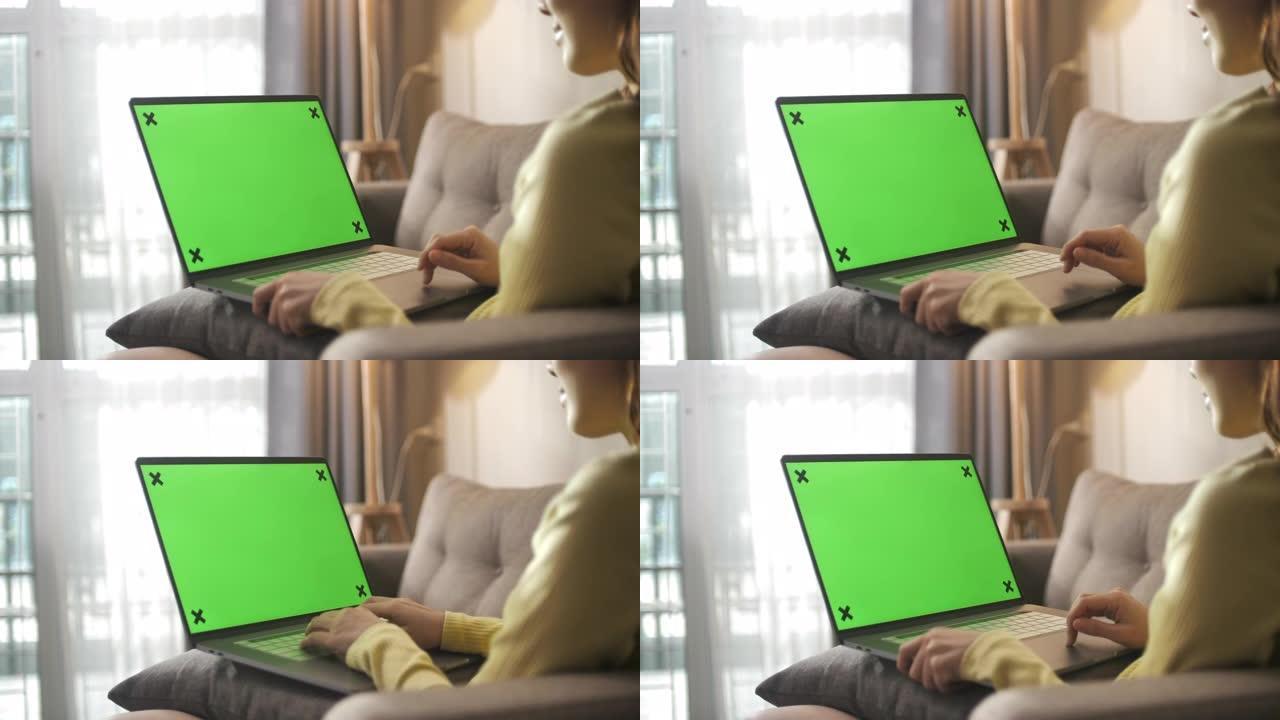 亚洲妇女坐在沙发上，在家中使用带有绿屏的笔记本电脑