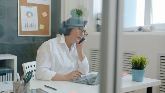 开朗的女人用笔记本电脑打字，然后用手机聊天，独自在办公室工作，微笑着