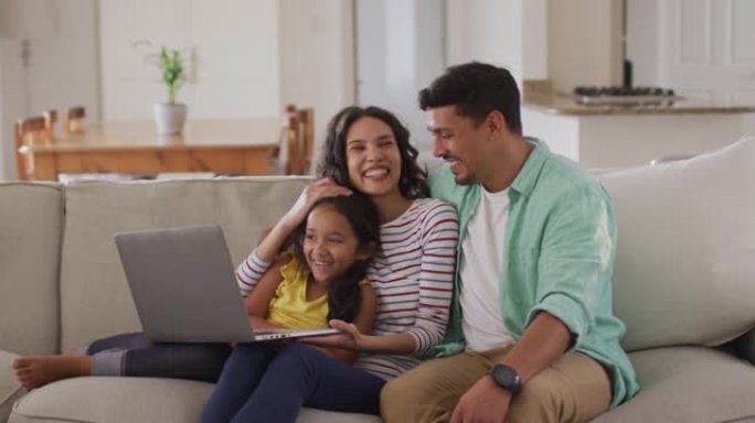 快乐的西班牙裔父母和女儿坐在沙发上使用笔记本电脑