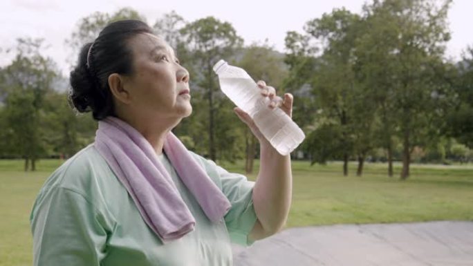 手持镜头: 亚洲高级女子早上在公园跑步后喝水。她拿着一瓶塑料水。做了瑜伽和太极运动后，她累了。医疗保