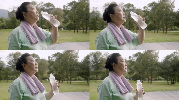 手持镜头: 亚洲高级女子早上在公园跑步后喝水。她拿着一瓶塑料水。做了瑜伽和太极运动后，她累了。医疗保