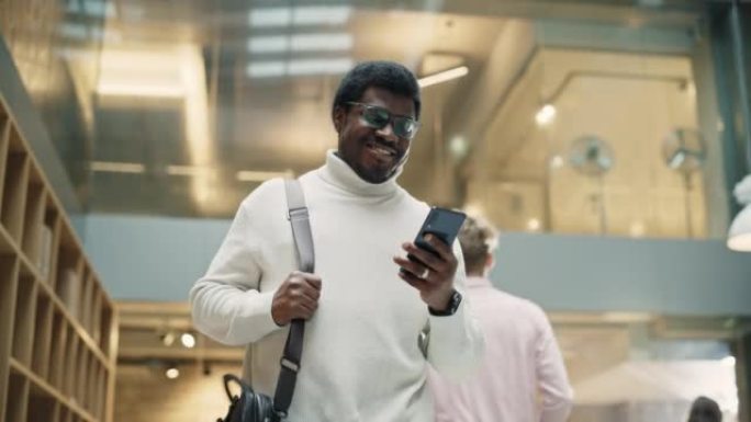 年轻黑人男子的肖像走进公司办公室走廊，用智能手机微笑着。成功的商人通过短信与同事交流。慢动作，低角度