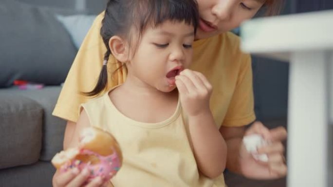 快乐快乐的亚洲家庭妈妈和蹒跚学步的女孩吃甜甜圈，玩得开心放松，在家里客厅的沙发上享受。