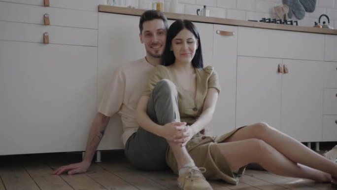 幸福的年轻夫妇坐在厨房的地板上微笑着看着相机的肖像
