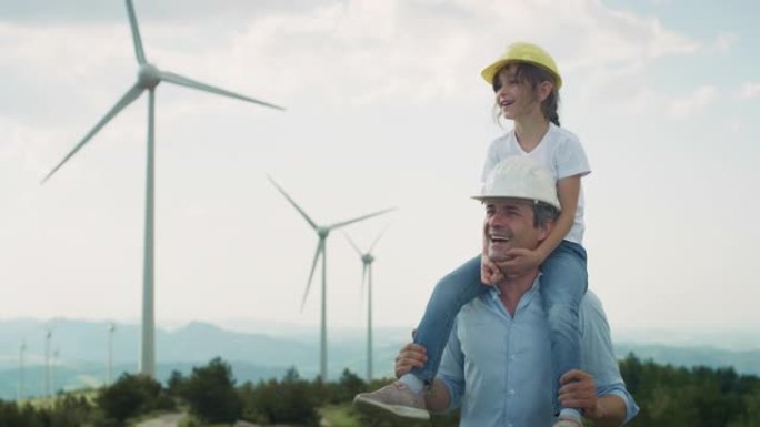 父亲和女儿戴着防护头盔，微笑着指着风电场，背景是风力涡轮机。为子孙后代和我们的孩子提供绿色能源和生态