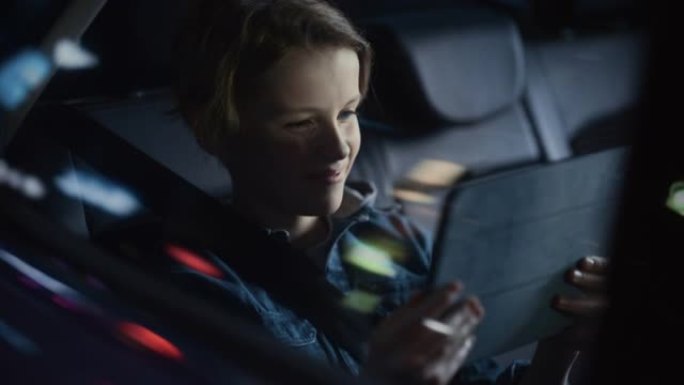 英俊的小男孩坐在汽车后座上，晚上通勤回家。乘客在带有霓虹灯的城市街道上乘坐出租车时，在平板电脑上观看