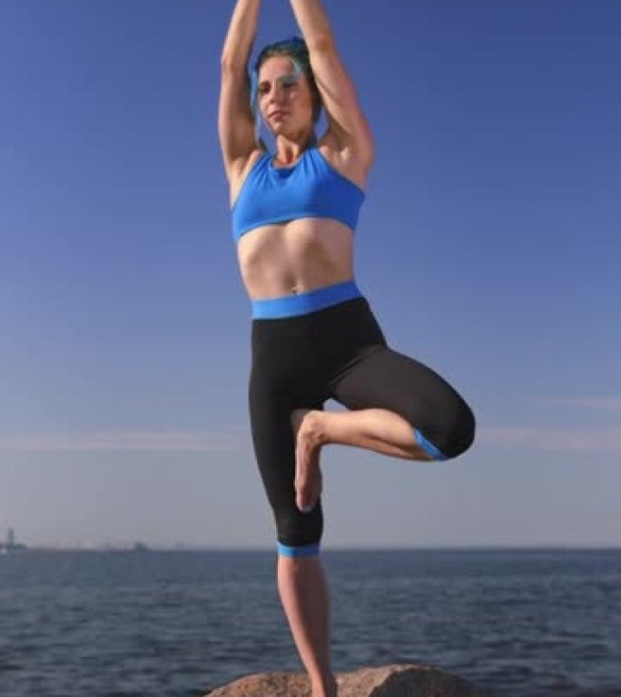 一位迷人的健美瑜伽老师，蓝色的头发穿着蓝黑色的瑜伽服，站在白天的天空和大海的背景下，她正在做树瑜伽姿