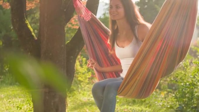快乐的年轻女子在后院树下的吊床上摇摆，微笑着