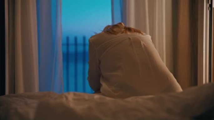 晚上，金发女人坐在酒店床的边缘，低着头，背景是大海的阳台