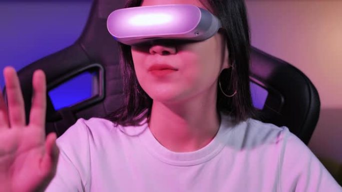 亚洲少女戴着虚拟现实耳机，坐在办公桌前打手势，晚上在彩色紫外线照明客厅中触摸空气。技术