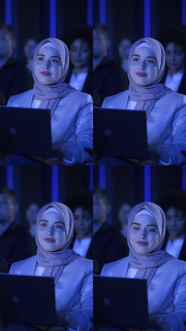 垂直屏幕: 阿拉伯女性坐在科技会议上黑暗拥挤的礼堂里。使用笔记本电脑的年轻穆斯林妇女。观看创新技术演