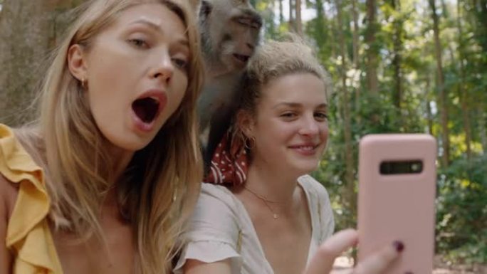 最好的朋友使用智能手机与猴子坐在肩膀上拍照两个女人在野生动物动物园与猴子合影，游客在巴厘岛旅行