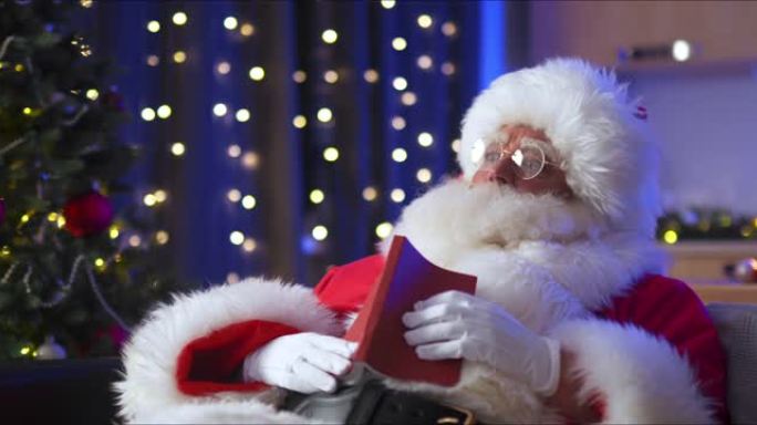 圣诞老人坐在装饰房间的沙发上看书，身后有一棵圣诞树
