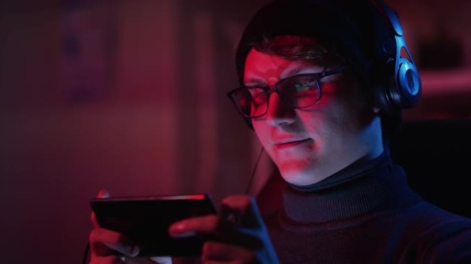 一位戴着无檐小便帽和眼镜的年轻人游戏玩家的电影镜头正在使用智能手机播放隔离在黑暗背景上的最新创新技术