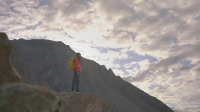 自由快乐的旅行者年轻女性穿着橙色的冬季夹克，背着黄色的背包站在山顶上，早晨可以看到晴朗的天空。种族多