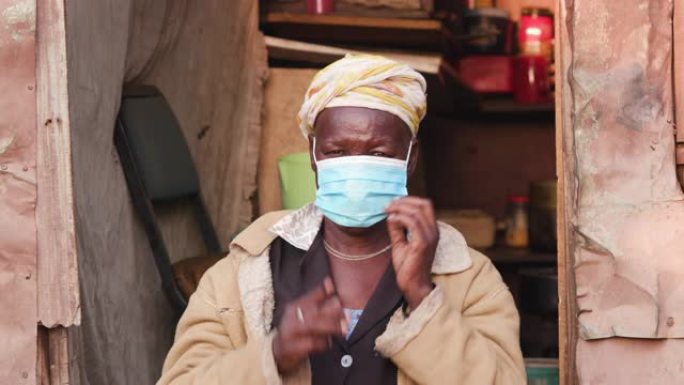 新型冠状病毒肺炎期间，一名非洲黑人老妇人在一个非正式定居点的铁皮小屋门口戴上防护口罩的特写肖像