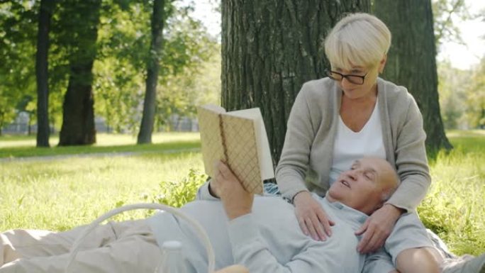 快乐的老年夫妇在公园的草坪上看书和大笑的慢动作肖像