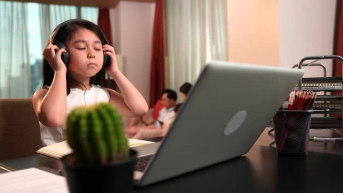 亚洲女孩正在通过笔记本电脑和耳机在线学习，一家人坐在后台的沙发上。社交距离，儿童教育，自学，电子学习