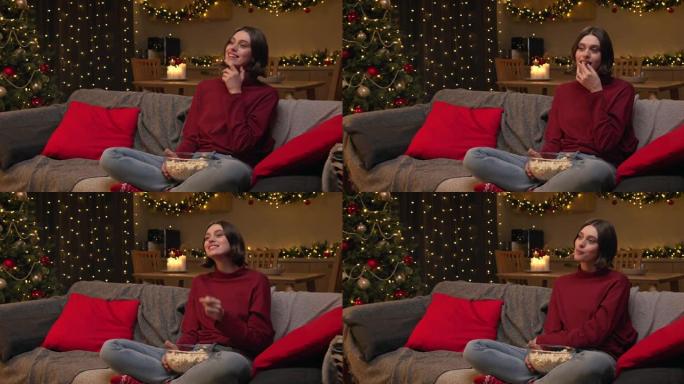 一个穿着红色毛衣的年轻美女坐在舒适的圣诞节装饰房间的沙发上，拿着一碗爆米花看电视