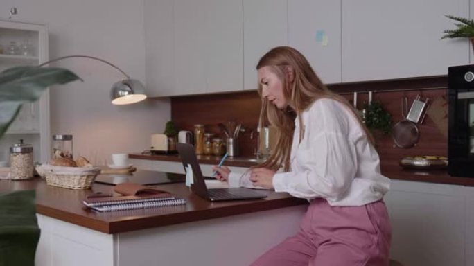 过度劳累的概念。严肃的金发白人女商人使用笔记本电脑，在家里厨房的桌子上做笔记。
