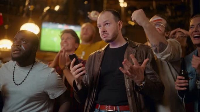 一个严肃的年轻人拿着智能手机的肖像，对足球比赛的体育赌注感到焦虑。看起来像一家普通的英国酒吧，穿着一