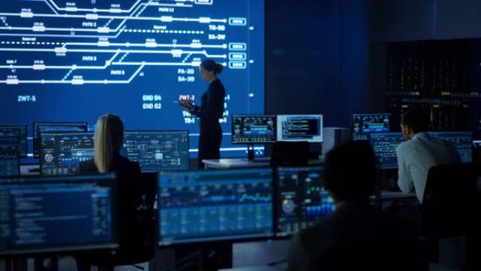 女性项目负责人使用数字平板电脑，看着大屏幕显示屏，显示基础设施信息图表和数据。电信控制监控室，工作人