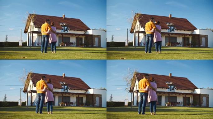 SLO MO夫妇站在草坪上拥抱，他们欣赏他们的新房子