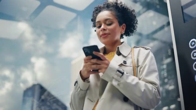 自信的黑人女性乘坐玻璃电梯到现代商务中心的办公室。成功的非洲裔美国经理在使用智能手机时微笑，写短信，