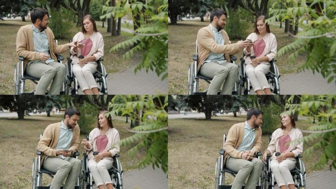 残疾男子在户外与残疾女孩交谈，拿着她的智能手机坐在轮椅上享受公园