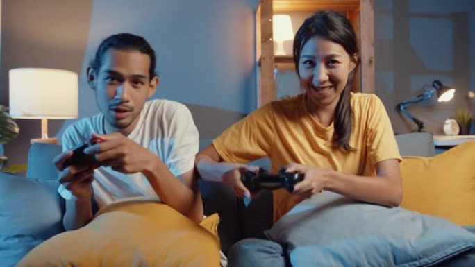 快乐亚洲年轻夫妇坐在沙发上使用操纵杆控制器玩视频游戏，晚上在客厅一起度过快乐的时光。