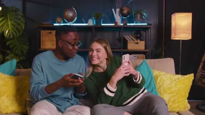 两个开朗的年轻快乐的多民族朋友使用手机在线购物，一起在家里的沙发上玩游戏。