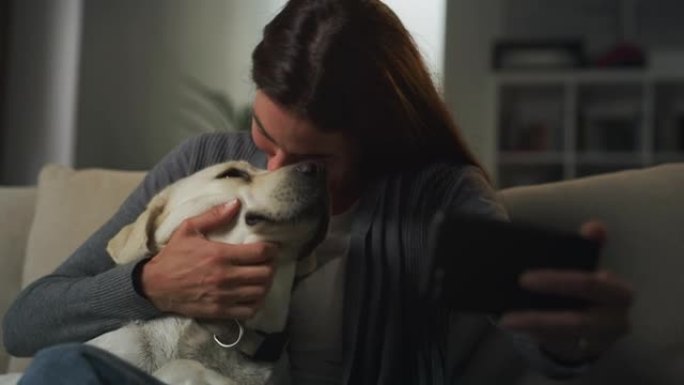 一名妇女与拉布拉多犬狗坐在沙发上的肖像，使用智能手机并进行视频通话。年轻女性在搬家后与家人和朋友保持