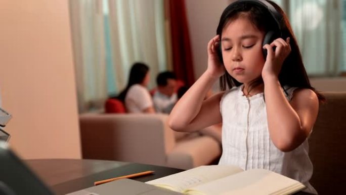 亚洲女孩在网上学习时通过笔记本电脑和耳机做笔记，家人坐在沙发上的背景。社交距离，儿童教育，自学，电子