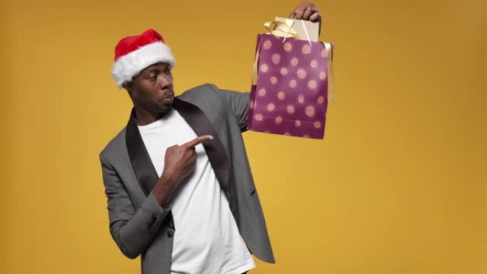 一个戴着圣诞帽和灰色夹克的黑皮肤男人举起双手，用带有礼品盒的包裹
