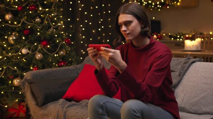 一个穿着红色毛衣和牛仔裤的棕色头发的年轻漂亮女人坐在一个舒适的圣诞节装饰的房间里，在智能手机上输入一