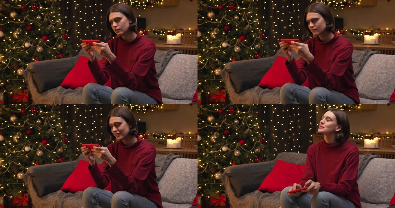 一个穿着红色毛衣和牛仔裤的棕色头发的年轻漂亮女人坐在一个舒适的圣诞节装饰的房间里，在智能手机上输入一