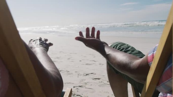 老年非裔美国夫妇坐在日光浴床上，在海滩上牵手