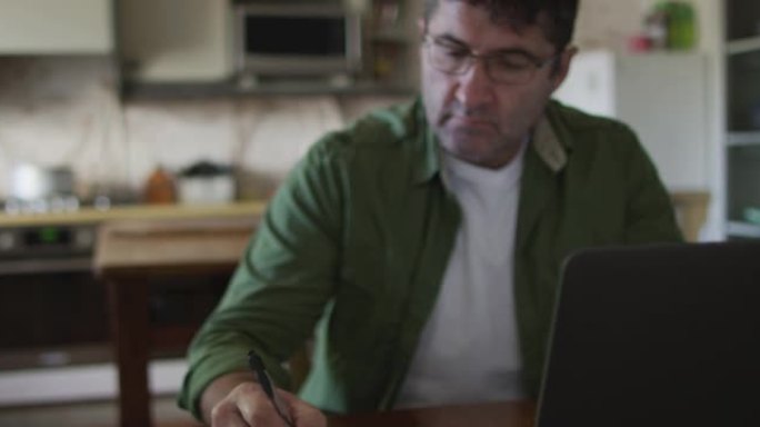 白人男子坐在餐桌旁，使用笔记本电脑并在家做笔记