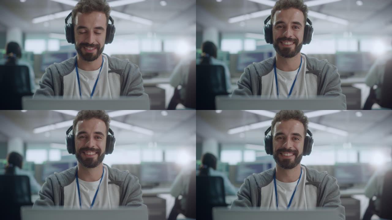 办公室: 快乐的IT程序员的肖像戴着耳机在台式计算机上工作，看着相机并微笑着。男软件工程师开发应用程