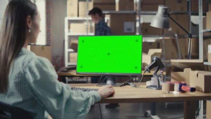 女性使用带有绿屏Chromakey模拟显示的台式计算机。两个小企业主拿着纸板箱在配送仓库工作。肩膀上