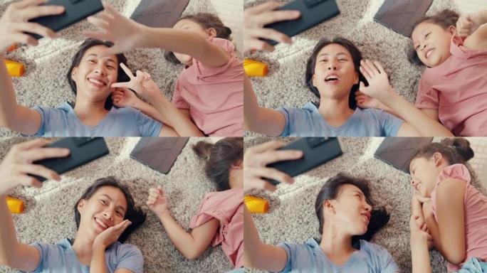 亚洲蹒跚学步的小女孩女儿与母亲使用智能手机在地毯地板上拍照或视频通话，在家里的客厅开心，开心。