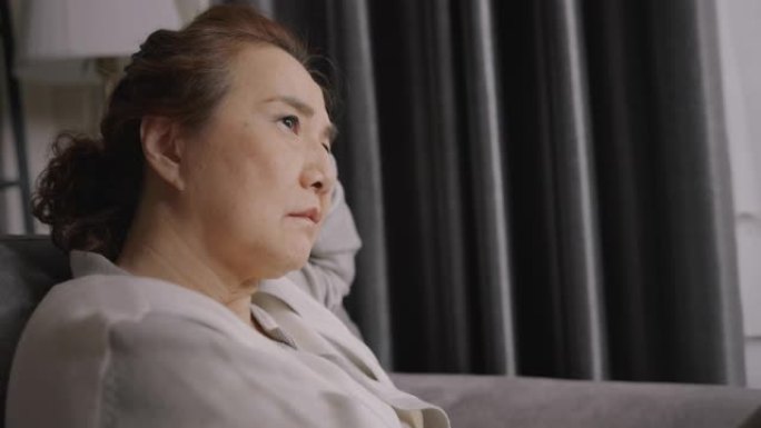 亚洲高级女性在家坐在客厅的沙发上时感到悲伤和担心自己的健康问题或生病