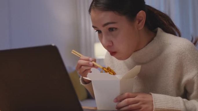 年轻迷人美丽的亚洲女性饥饿吃方便面中国食品盒拿走满嘴看电脑笔记本在家里忙碌的工作深夜多任务不健康的一