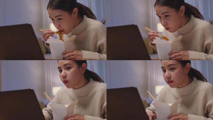年轻迷人美丽的亚洲女性饥饿吃方便面中国食品盒拿走满嘴看电脑笔记本在家里忙碌的工作深夜多任务不健康的一
