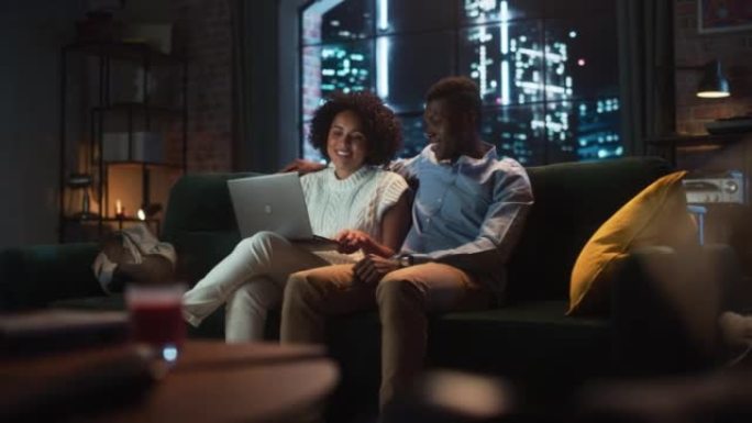 年轻的黑人夫妇坐在舒适时尚的公寓里的沙发上，使用笔记本电脑。男女朋友进行网上购物，使用社交媒体，观看