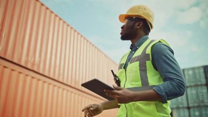一位穿着黄色安全帽和安全背心的英俊非洲裔美国黑人工业工程师在平板电脑上工作的微笑肖像。集装箱码头的检