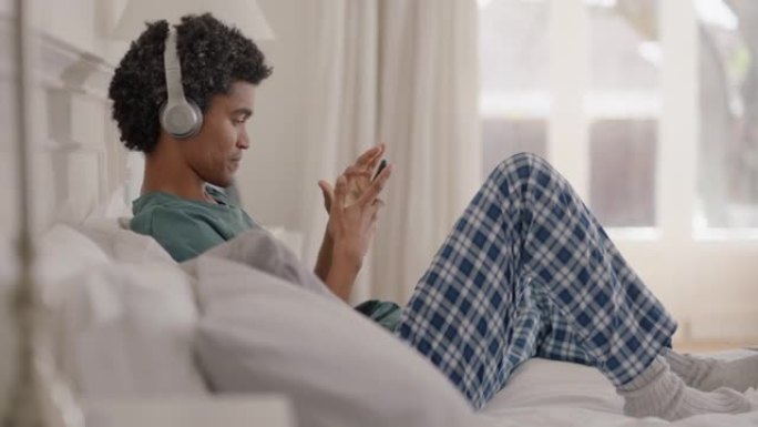 年轻的混血男子使用智能手机听音乐戴着耳机浏览在线放松床上分享生活方式在社交媒体上享受周末早上