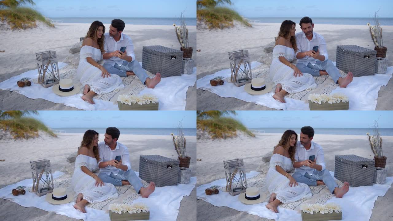 幸福的夫妇在海滩上野餐，一边看智能手机上的社交媒体一边放松和交谈