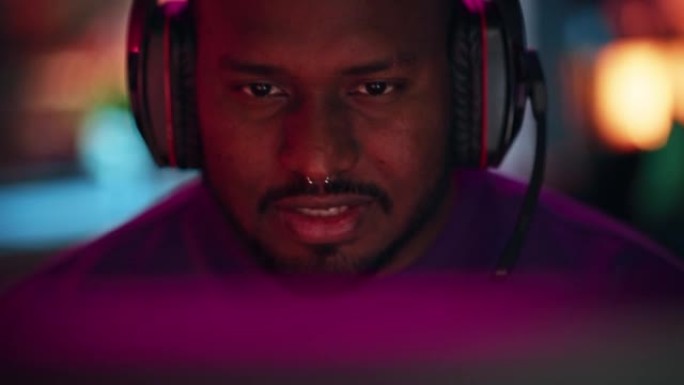 一个非洲裔美国时尚年轻人的特写肖像，他的鼻环在晚上在家玩在线电脑视频游戏。玩家在与耳机交谈时与队友讨
